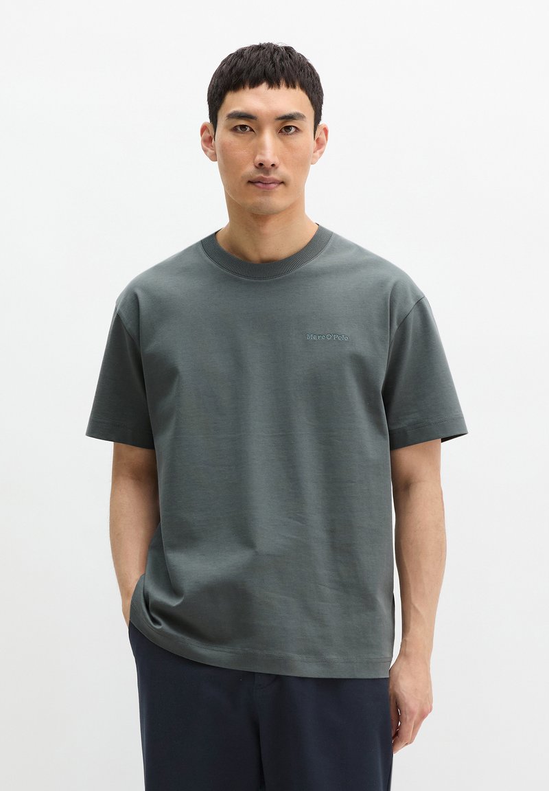 Homme aux cheveux noirs raides portant un t-shirt gris Marc O’Polo et un pantalon sombre, debout devant un fond blanc uni.