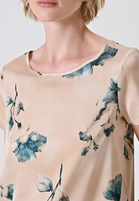 Top floreale beige realizzato in tessuto morbido. Presenta un collo rotondo e motivi floreali blu-verdi. Maniche corte e vestibilità ampia.