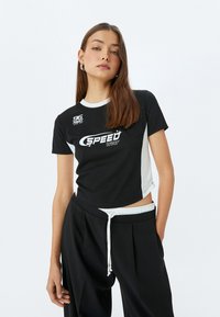 Sort og hvid crop t-shirt med korte ærmer, der har en rund halsudskæring og "SPEED" logoet trykt på forsiden. Sammen med løse sorte bukser.