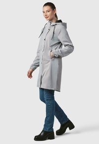 Lys grå vandtæt jakke med hætte, der har elastiske manchetter, trykknapper og to sidelommer, båret over blå jeans og sorte platformstøvler.