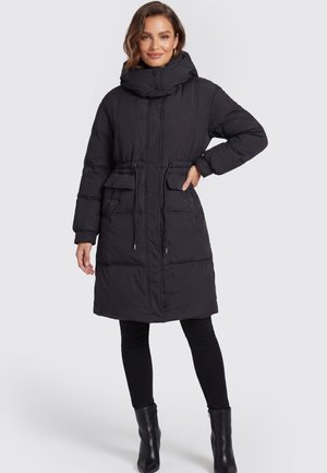 Femme portant un long manteau d'hiver noir matelassé à col montant, poches plaquées, taille à cordon, leggings noirs et bottines noires.