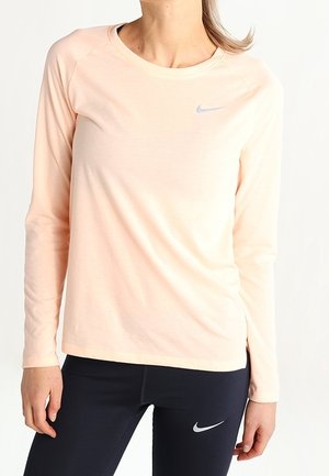 Žena oblečená v světle růžovém tričku s dlouhým rukávem značky Nike a černých legínách značky Nike, stojící před čistě bílým pozadím.