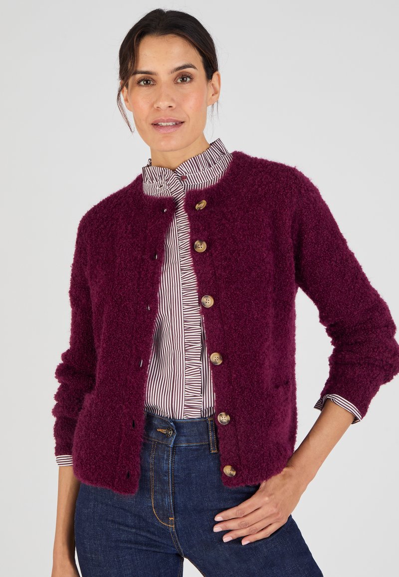 Cardigan borgogna e morbido con collo rotondo, dotato di cinque grandi bottoni sul davanti e polsini a coste. Indossato sopra una camicia a righe.