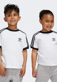 Biele tričká Adidas s čiernymi rebrovanými goliermi a tromi čiernymi pásmi na rukávoch, nosené s šedými teplákmi. Logo na hrudi.