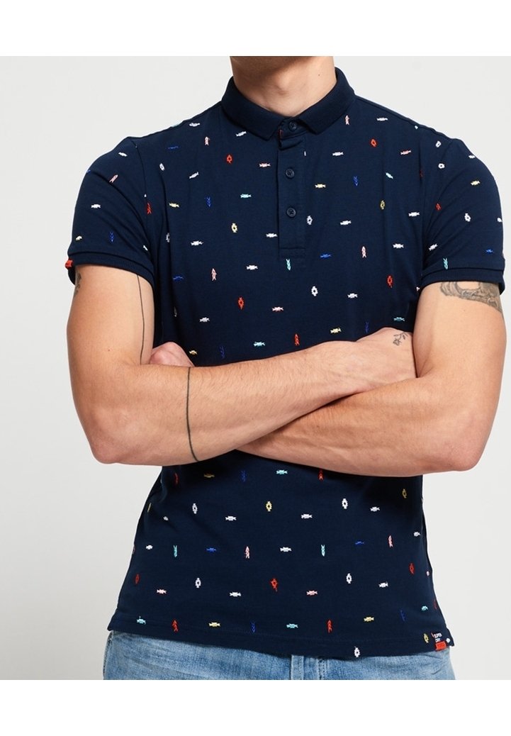 Homme les bras croisés portant un polo bleu marine avec de petits motifs brodés colorés et un jean bleu clair.