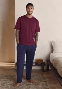 Bordowa koszulka z kieszenią i granatowe spodnie, oba wykonane z miękkiej tkaniny. Model stoi boso na teksturowanym dywanie w neutralnym pomieszczeniu.