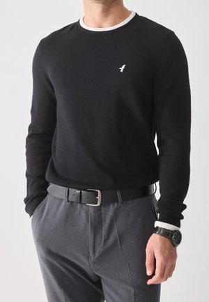 Mann trägt schwarzen Langarmpullover mit weißem Kragen und kleinem Vogel-Logo, graue Hose, schwarzer Gürtel und schwarze Armbanduhr, Hände in den Taschen.