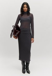 Langes, tailliertes Kleid mit hohem Kragen, aus durchsichtiger schwarzer Stoff mit weißen Punkten. Kombiniert mit knielangen schwarzen Stiefeln und einer burgunderfarbenen Tasche.