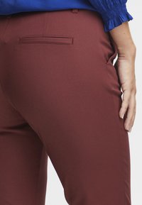 Bordó kalhoty se hladkým pružným materiálem a zadní kapsou. Detailní pohled z boku, ruka opřená o bok.