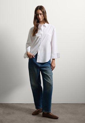 White button-up shirt with long sleeves, paired with blue loose-fitting jeans and brown flat shoes. The shirt has a single breast pocket.