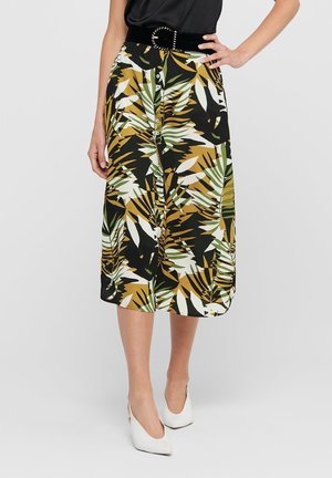Kobieta ubrana w spódnicę na guziki z tropikalnym wzorem liści w kolorach zielonym, białym, musztardowym i czarnym, w połączeniu z białymi szpilkami i czarnym paskiem.