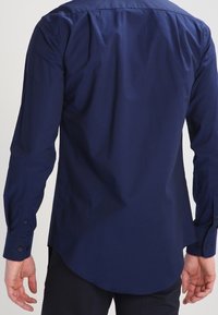 Man in een donkerblauwe overhemd met lange mouwen en zwarte broek, gezien van achteren met ontspannen armen langs zijn zij.