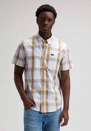 Junger Mann in einem kurzärmligen, karierten Hemd mit beigen, weißen und blauen Streifen und blauen Jeans, der vor einem schlichten Hintergrund steht.