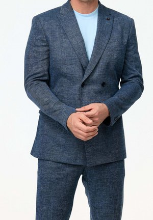 Hombre con traje azul de doble botonadura texturizado, pantalones a juego y camiseta de cuello redondo azul claro, manos entrelazadas al frente.