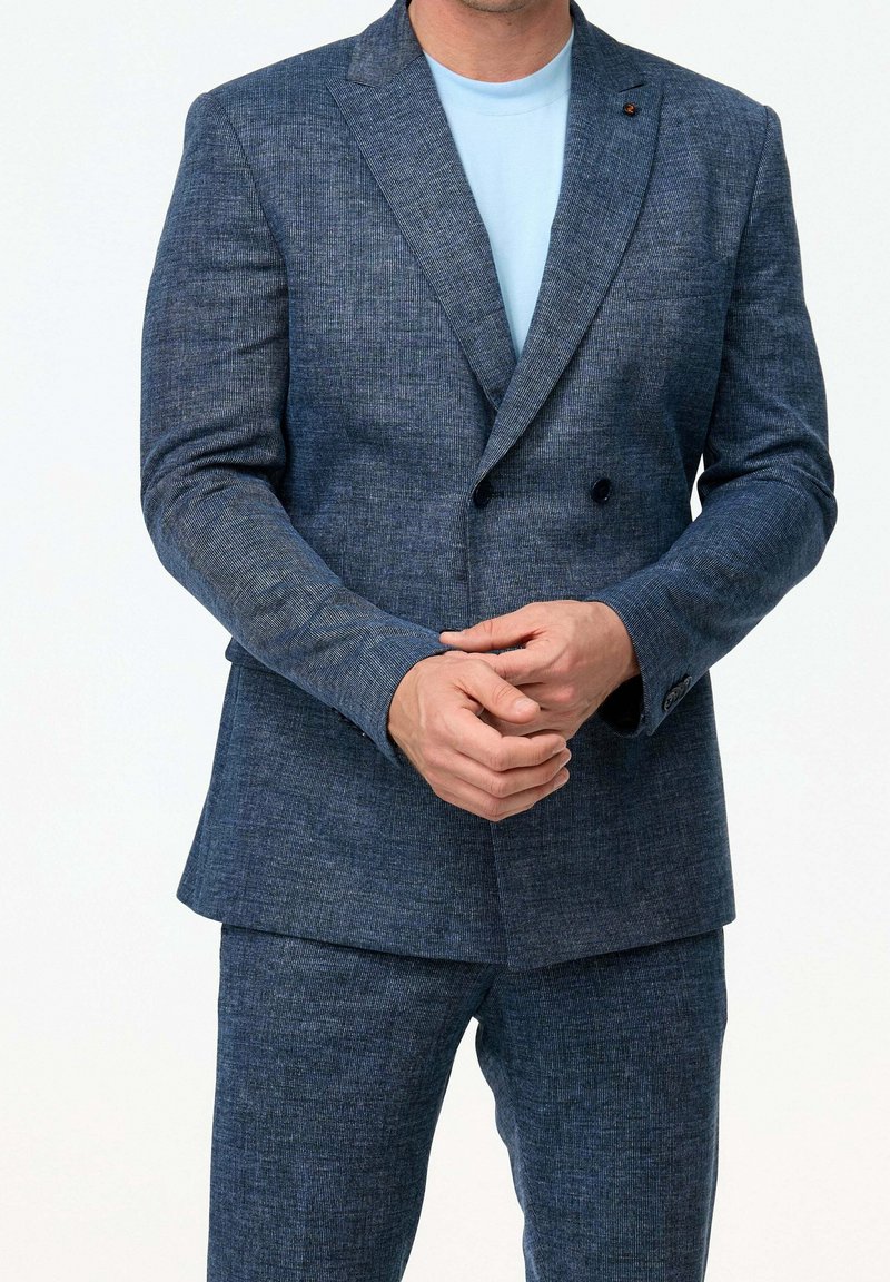 Homme portant un costume bleu texturé à double boutonnage avec un pantalon assorti et un t-shirt bleu clair à col rond, mains jointes devant.