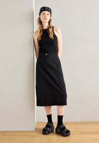 Débardeur noir et jupe noire longueur genou avec poches. Des chaussures à plateforme noires et des chaussettes complètent la tenue. Design simple et minimaliste.