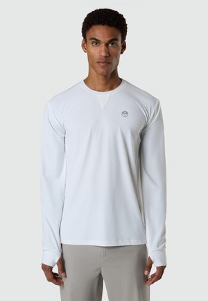 Mann trägt ein langärmeliges weißes Rundhalshemd mit einem kleinen, runden Kompass-Logo auf der Brust, steht vor einem einfarbigen Hintergrund.