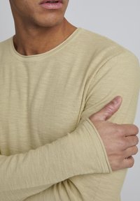 Homme vêtu d'une chemise beige à manches longues, le pouce accroché sous le bras, faisant légèrement face à gauche sur un fond clair et uni.