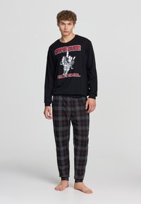 Maglietta nera a maniche lunghe con stampa grafica di una mano e controller di gioco; abbinata a pantaloni morbidi e rilassati in plaid grigio e nero.