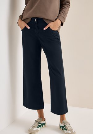 Personne portant un pantalon large court bleu marine, un haut marron à manches longues et des baskets blanches avec des accents verts et beiges, debout de manière détendue.