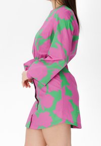 Woman wearing a fitted long-sleeve mini dress with a bold green floral pattern on a pink background, standing against a white backdrop.