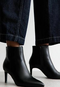 Botas de cuero negras hasta el tobillo con puntas afiladas y tacones delgados. Cuentan con una cremallera lateral y una textura suave. Combinadas con pantalones de mezclilla oscuros de pierna ancha.