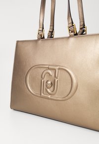 Borsa in bronzo metallico con texture liscia, caratterizzata da un logo stampato in rilievo e attacchi per tracolla piatta con hardware dorato.