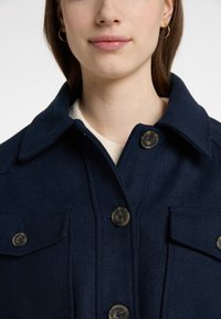 Jachetă dintr-un amestec de lână de culoare bleumarin, cu un guler structurat, două buzunare pe piept și închideri cu butoane, având butoane rotunde, texturate.