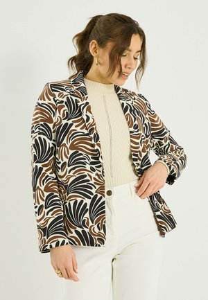 Femme portant un blazer à motifs de formes de feuilles marron, noir et crème sur un pull crème côtelé et un pantalon blanc, en train d'ajuster le blazer.