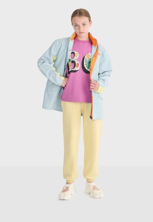Young person wearing a light blue jacket with orange lining, purple floral "BO" shirt, yellow pants, and white split-toe shoes, standing on white background.