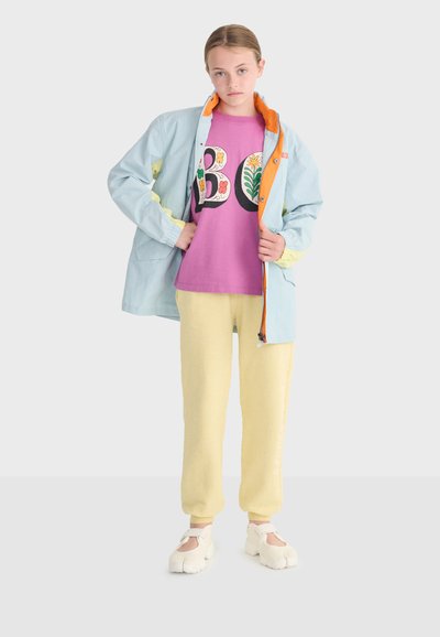 Jeune personne portant une veste bleu clair avec une doublure orange, un t-shirt violet à fleurs "BO", un pantalon jaune et des chaussures à orteils fendus blanches, debout sur un fond blanc.