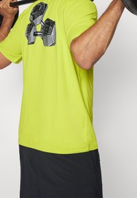 Persona sosteniendo una barra sobre los hombros, vistiendo una camiseta verde brillante con un gráfico de mancuerna negra y pantalones cortos negros.