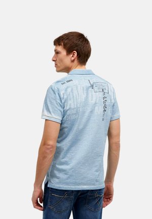 Mann in hellblauem grafischem Poloshirt und dunklen Jeans, von hinten gesehen, mit Text und Design auf dem oberen Rücken des Shirts.