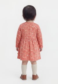 Copil mic stând cu fața spre departe, purtând o rochie corală cu flori și mâneci lungi, șosete crem până la genunchi și pantofi maro pe un fundal alb.