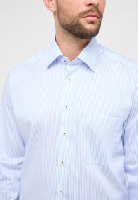 Chemise à rayures bleu clair en tissu lisse. Elle présente un col pointu, une patte de boutonnage et une poche poitrine unique.