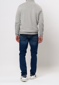 Man wearing light gray sweater, dark blue jeans, and white sneakers with navy and red details, standing facing away on a plain background.