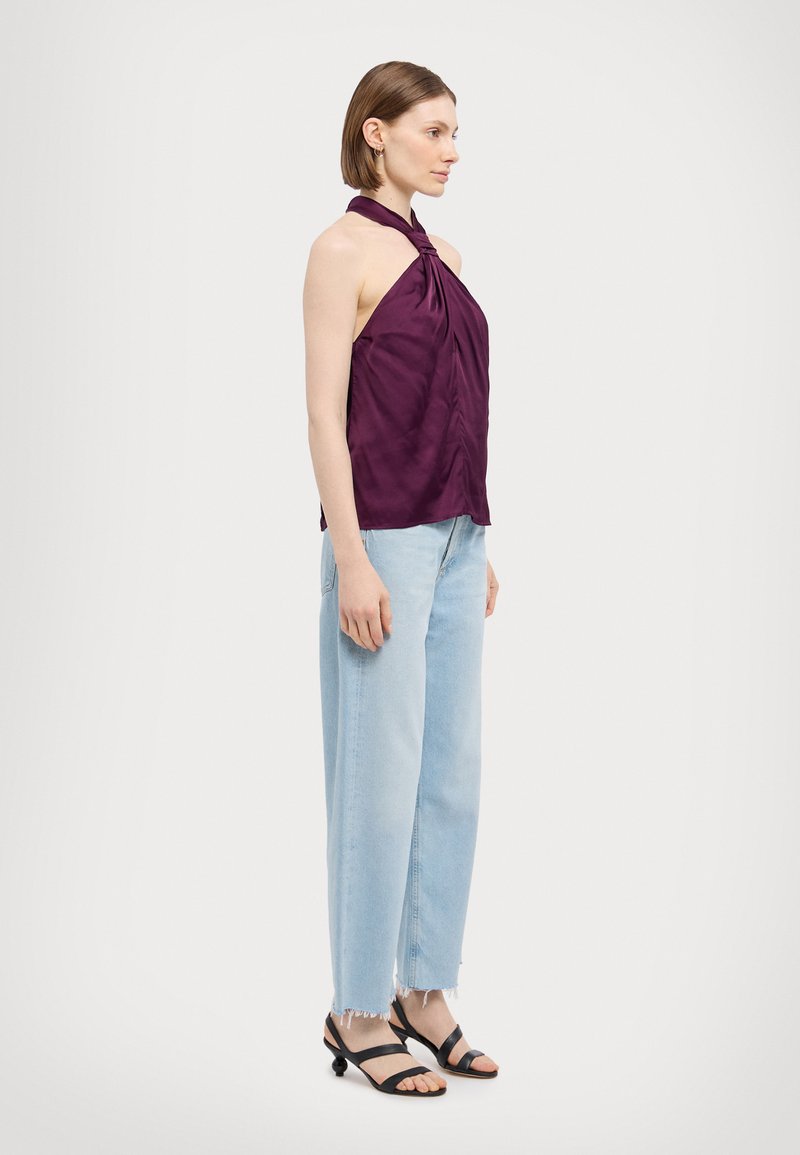 Top halter viola con dettaglio nodo, caratterizzato da una superficie liscia. Abbinato a jeans azzurri chiari a vita alta e sandali neri con tacco basso.
