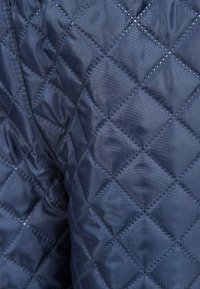 Laikkuvalkoinen tikkikangas, jossa on timanttimuotoilu, joka esittelee sileän tekstuurin ja näkyvät ompeleet. Ei muita yksityiskohtia tai koristeita.