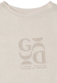 T-shirt beige à col rond avec des formes abstraites et le texte « TIMES AHEAD FOLLOW your DREAMS » imprimé en tons atténués sur le devant.