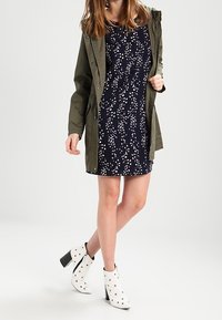 Olivgrön parka med tryckknappar över en marinblå blommig klänning; vita ankelskotter med nitar; smidig, modern design och silhuett.