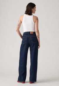 Blugi din denim albastru închis cu croială largă, talie înaltă, buzunare posterioare vizibile cu cusături curbate și o etichetă de brand din piele.