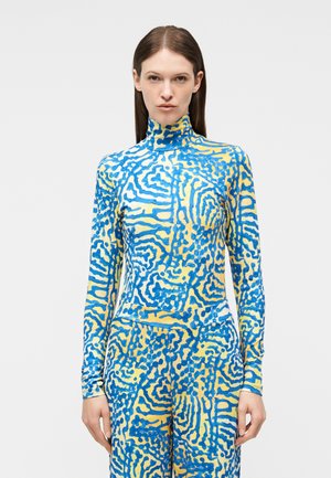 Γυναίκα που φοράει μακρυμάνικο τοπ με κολάρο σε κίτρινο και μπλε με αφαίρετο μοτίβο και παντελόνι που ταιριάζει, στέκεται μπροστά σε απλό φόντο.