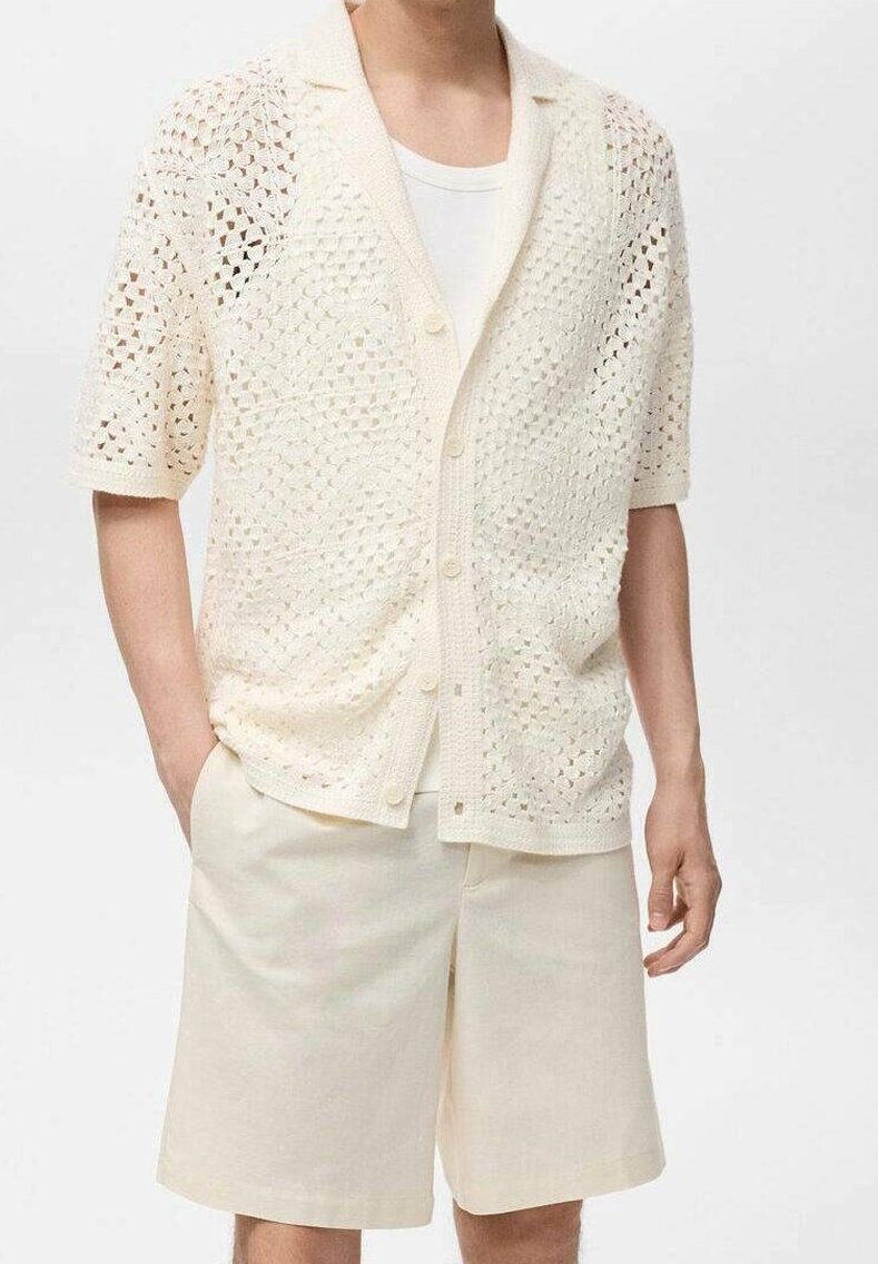 Homme portant un cardigan crème en crochet à manches courtes boutonné sur un t-shirt blanc avec un short crème à hauteur des genoux, mains relâchées sur les côtés.