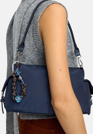 Granatowa torba na ramię z regulowanym paskiem i ozdobą w postaci supełkowanego breloczka z liny, noszona na ramieniu wraz z szarym bezrękawnikiem z dzianiny.