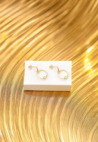 Guldörhängen med blommig stud, med en slät rund form och en liten kula som accent vid botten, uppvisade på en vit låda.