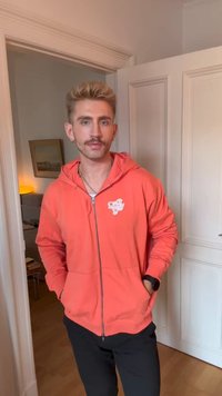 Man with mustache and short blonde hair, wearing an orange zip-up hoodie and black pants, standing in a doorway indoors.