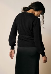 Cardigan noir en tricot avec une finition texturée et des manches bouffantes, doté d'une taille ceinturée et d'une coupe décontractée, associé à une jupe noire.