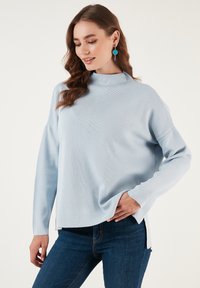 Világoskék kötött pulóver magas gallérral, hosszú ujjal és oldalsó hasítékokkal. Sötét farmernadrággal párosítva. Simogatós textúra finom bordázással.