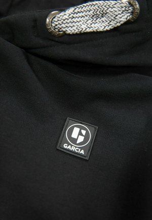 Schwarzer Stoff mit glatter Textur, der ein quadratisches Etikett mit dem "GARCI"-Logo und eine geflochtene Kordel mit metallischem Akzent zeigt.