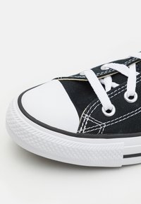 Svart kanvas sneakers med vit gummikappa och sula. Har vita skosnören och sömmar, metallöglor och en svart-och-vit randig kant.